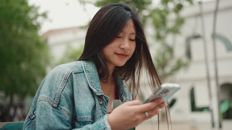 Mujer-Asiática-Revisando-Su-Teléfono-Mientras-Está-Sentada-En-Un-Banco-En-El-Parque.