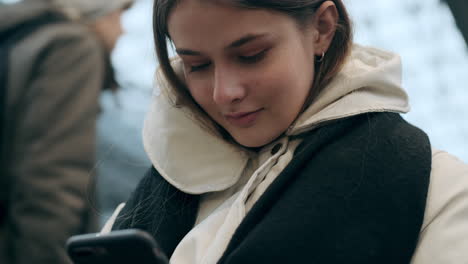 Caucasian-girl-in-down-jacket-with-scarf-dreamily-using-cellphone-sitting-at-underground-station.