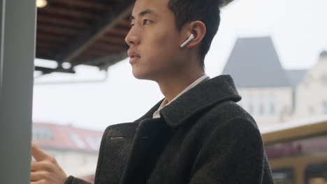 Young-Asian-businessman-in-coat-with-wireless-earphones-thoughtfully-watching-public-transport-route-waiting-on-street.