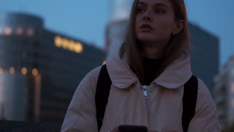 Medium-shot-of-Caucasian-girl-thoughtfully-looking-aside-using-cellphone-on-evening-street-at-dusk.