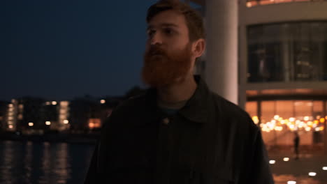 Tracking-around-shot-of-young-handsome-redhead-bearded-man-thoughtfully-walking-through-city-street-at-night.