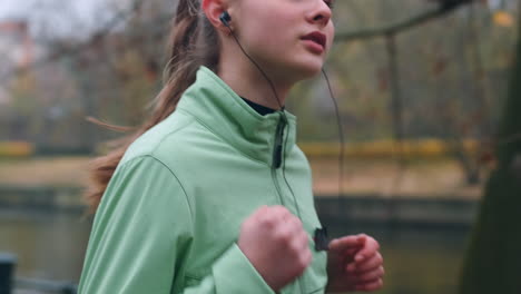 Sporty-Caucasian-girl-in-earphones-running-in-autumn-city-park-along-river.