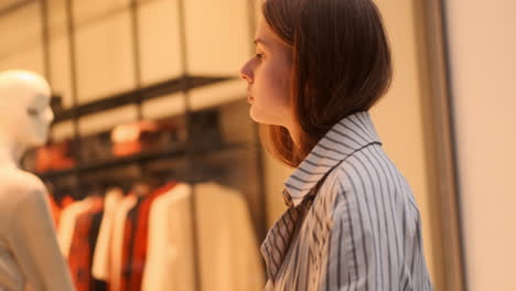 Side-view-of-stylish-Caucasian-girl-in-trench-coat-thoughtfully-examining-shop-window-on-city-street.