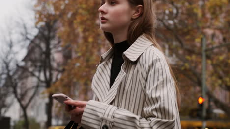 Tracking-in-shot-of-Caucasian-girl-in-trench-coat-with-cellphone-confidently-walking-through-city-street.