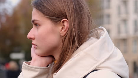 Side-view-of-Caucasian-girl-in-down-jacket-leaning-on-hand-thoughtfully-looking-away-on-city-street.