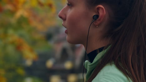 Tracking-shot-of-sporty-girl-thoughtfully-wearing-earphones-preparing-for-running-in-autumn-city-park.