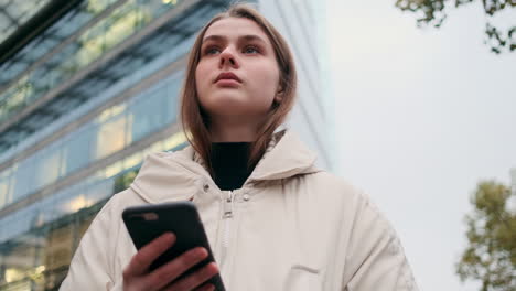 Caucasian-girl-in-down-jacket-with-cellphone-thoughtfully-waiting-on-city-street.