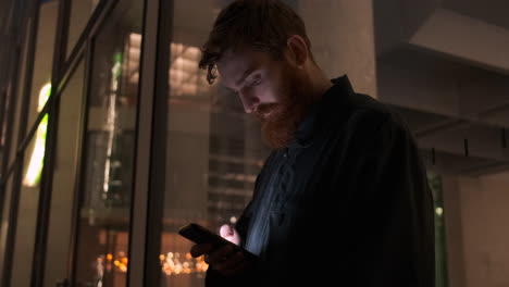 Young-handsome-casual-bearded-man-intently-using-cellphone-on-city-street-at-night-outside-business-center.