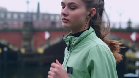 Sporty-Caucasian-girl-in-earphones-confidently-running-in-autumn-city-park-on-riverside.