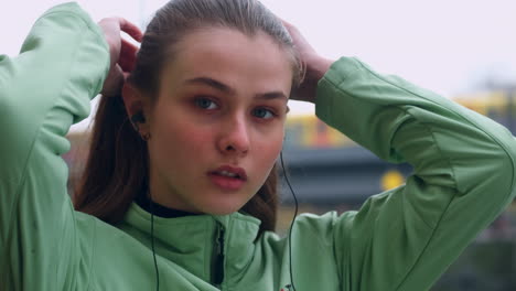 Portrait-of-sporty-Caucasian-girl-in-earphones-tying-hair-in-tail-intently-looking-in-camera-in-city-park.