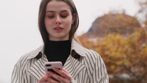 Tracking-shot-of-Caucasian-girl-in-trench-coat-dreamily-using-cellphone-on-city-street.