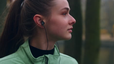 Sporty-Caucasian-girl-in-earphones-dreamily-walking-after-running-in-autumn-city-park.