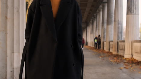 Tracking-shot-of-stylish-Caucasian-girl-in-coat-confidently-walking-through-archway-in-city-park.