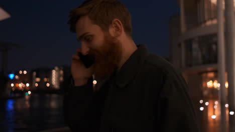 Tracking-shot-of-young-bearded-man-joyfully-talking-on-cellphone-walking-through-city-street-at-night.