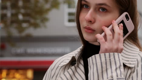 Chica-Caucásica-Con-Gabardina-Hablando-Pensativamente-Por-Teléfono-Celular-En-La-Calle-De-La-Ciudad.