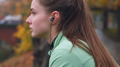 Verfolgungsaufnahme-Eines-Sportlichen-Kaukasischen-Mädchens-Mit-Kopfhörern,-Das-Sich-Nach-Einem-Lauf-Im-Stadtpark-In-Der-Nähe-Des-Flusses-Ausruht.