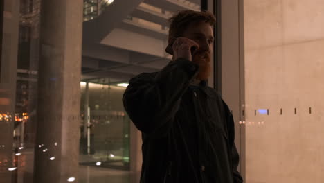 Young-bearded-man-happily-talking-on-cellphone-walking-through-city-street-outside-business-center-at-night.