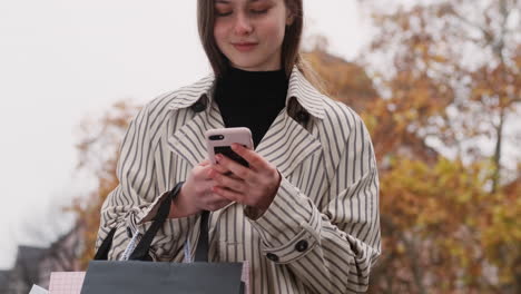 Portrait-of-stylish-Caucasian-girl-in-trench-coat-happily-using-cellphone-on-city-street.