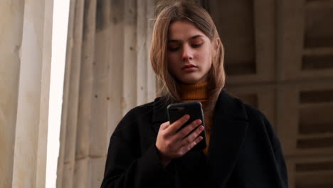 Caucasian-girl-in-coat-intently-using-cellphone-walking-through-archway-in-city-park.