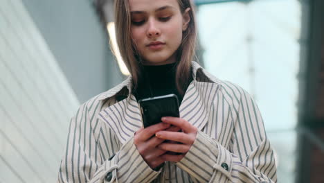 Portrait-of-stylish-Caucasian-girl-using-cellphone-thoughtfully-looking-in-camera-on-stairs-at-subway-station.