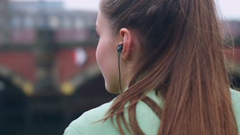 Tracking-around-shot-of-sporty-Caucasian-girl-in-earphones-dreamily-resting-after-running-in-city-park-on-riverside.