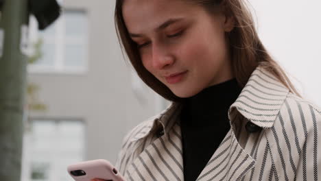 Close-up-shot-of-Caucasian-girl-in-trench-coat-happily-using-cellphone-waiting-on-city-street.