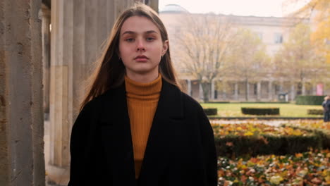 Portrait-tracking-shot-of-stylish-Caucasian-girl-in-coat-intently-looking-in-camera-confidently-walking-through-autumn-city-park.