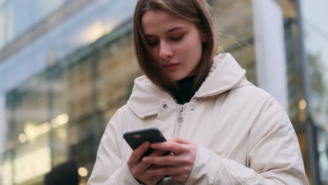 Portrait-of-Caucasian-girl-in-down-jacket-happily-using-cellphone-on-city-street.