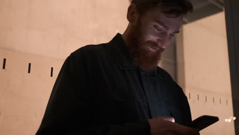 Tracking-shot-of-young-smiling-bearded-man-joyfully-using-cellphone-on-city-street-outside-business-center-at-night.