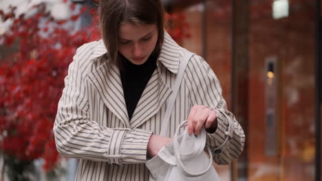 Stylish-girl-in-trench-coat-looking-something-in-woman-bag-on-city-street.