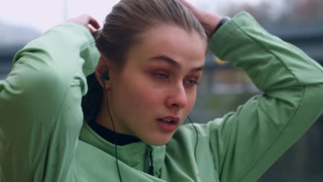 Portrait-of-sporty-Caucasian-girl-in-earphones-tying-hair-in-tail-and-start-running-in-city-park.