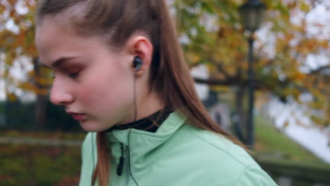 Sporty-Caucasian-girl-wearing-earphones-preparing-for-running-in-beautiful-autumn-city-park.