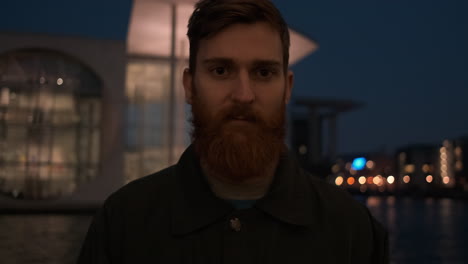 Portrait-shot-of-redhead-bearded-man-intently-looking-in-camera-on-city-street-at-night-on-riverside.