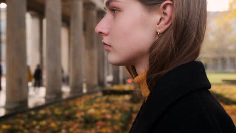 Side-view-of-Caucasian-girl-in-coat-confidently-walking-through-archway-in-city-park-at-sunset.