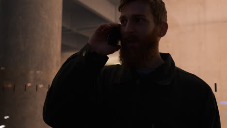 Tracking-shot-of-young-attractive-bearded-man-talking-on-cellphone-on-city-street-over-business-center-at-night.