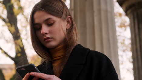 Caucasian-girl-in-coat-thoughtfully-using-cellphone-walking-outdoor-at-autumn.