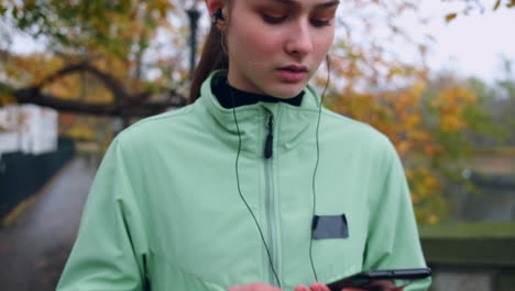 Tracking-out-shot-of-sporty-Caucasian-girl-wearing-earphones-preparing-for-running-in-autumn-city-park.