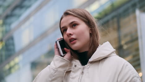 Caucasian-girl-in-down-jacket-talking-on-cellphone-thoughtfully-looking-round-on-city-street.