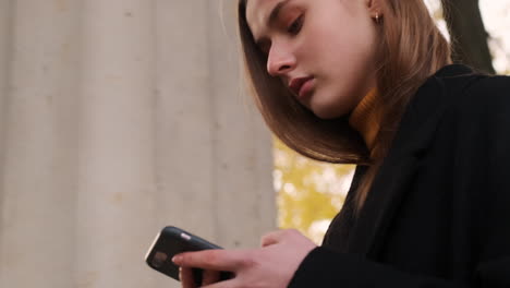 Chica-Caucásica-Con-Abrigo-Usando-Atentamente-Su-Teléfono-Celular-Caminando-Por-El-Parque-De-Otoño.