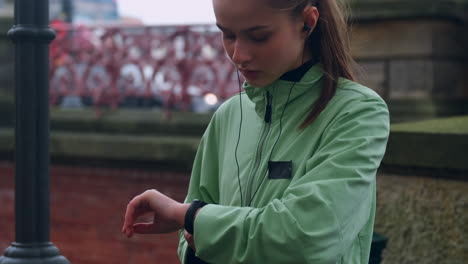 Seguimiento-De-Una-Chica-Deportista-Con-Auriculares-Preparándose-Pensativamente-Para-Correr-En-Un-Parque-De-La-Ciudad-Junto-Al-Río.