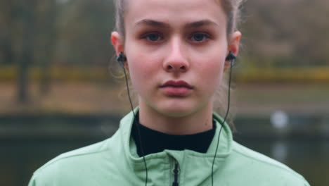 Portrait-of-sporty-Caucasian-girl-in-earphones-confidently-looking-in-camera-after-running-in-autumn-city-park-on-riverside.