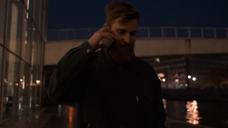 Tracking-around-shot-of-young-bearded-man-talking-on-cellphone-confidently-walking-through-city-street-at-night.