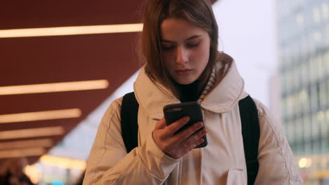 Caucasian-girl-in-down-jacket-with-backpack-intently-using-cellphone-on-city-street.