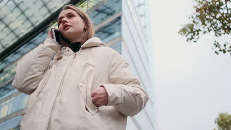 Caucasian-girl-in-down-jacket-talking-on-cellphone-waiting-someone-on-city-street.