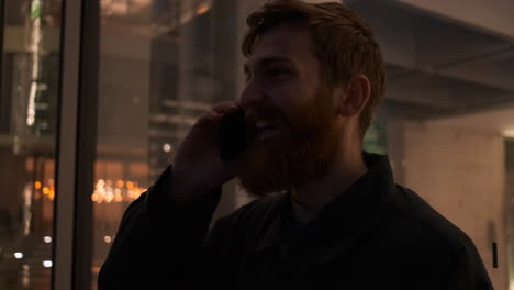 Tracking-around-shot-of-young-cheerful-redhead-bearded-man-happily-talking-on-cellphone-on-city-street-at-night.