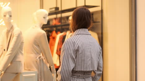 Back-view-of-stylish-Caucasian-girl-in-trench-coat-dreamily-examining-shop-window-on-city-street.