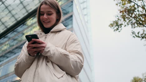 Portrait-of-casual-girl-in-down-jacket-happily-using-cellphone-waiting-on-city-street.