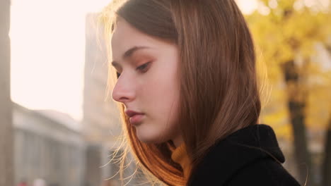 Side-view-of-Caucasian-girl-in-coat-dreamily-walking-on-city-street-at-sunset.