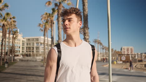 Tracking-shot-of-young-smiling-sporty-man-with-backpack-happily-walking-through-seaside