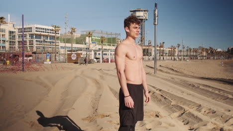 Tracking-in-shot-of-attractive-fit-guy-confidently-looking-in-camera-after-workout-on-beach